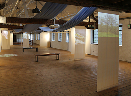A view of the whole exhibition in a mill building, with fabric screens and blue fabric woven through the beams in the ceiling