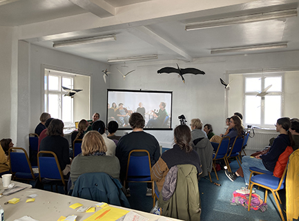 A room full of 16 people sitting on blue chairs, all facing the corner where there is a screen with a video projection on it