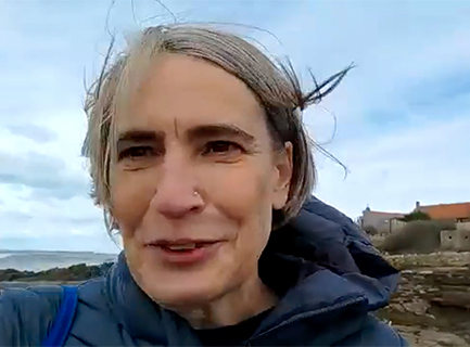 A smiling and talking woman with short grey hair wearing a grey hooded jacket