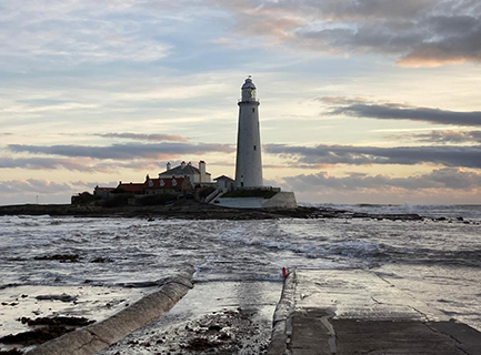 A lighthouse on an island with two other buildings, the sea covers the causeway, and the sun is setting to the right of the image